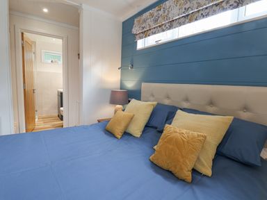 A bedroom with a blue bedspread yellow pillows a beige headboard a bedside lamp and a view into an adjoining bathroom at Hideaway Lodge in Runswick Bay
