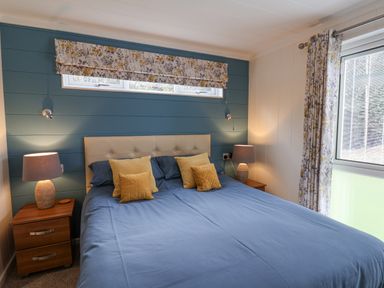 A bedroom with a double bed with blue bedding and yellow cushions two bedside tables with lamps and floral curtains at Hideaway Lodge in Runswick Bay