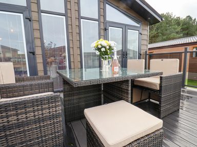 An outdoor patio with a glass table surrounded by wicker chairs with beige cushions and a vase of flowers on the table at Hideaway Lodge in Runswick Bay