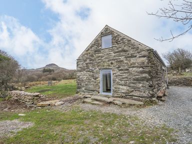 A stone building with two windows in a rural area with grass trees and a hill in the background at Hen Beudy in Porthmadog