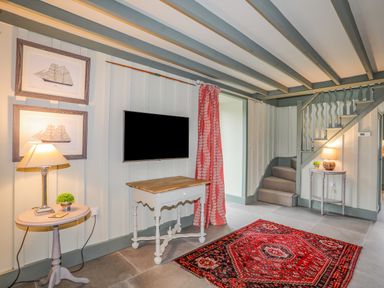 A room with a staircase two small tables with lamps a wall-mounted tv two framed ship pictures and a red patterned rug at Hen Beudy in Porthmadog