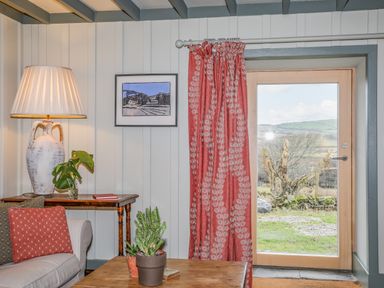 A corner of a living room with a sofa a lamp on a wooden side table framed art on the wall a red curtain and a glass door showing an outdoor view at Hen Beudy in Porthmadog
