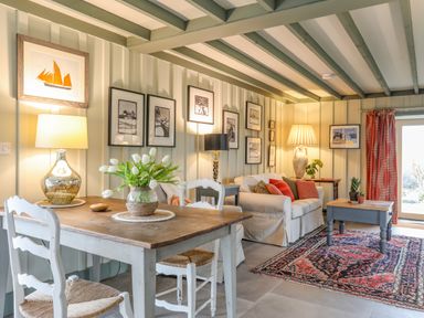 A living room with a wooden dining table and chairs white sofa table lamps framed pictures and a patterned rug at Hen Beudy in Porthmadog