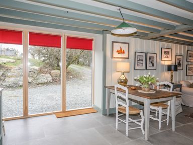 A dining area with a wooden table and two chairs next to large windows with red blinds and wall decorations at Hen Beudy in Porthmadog