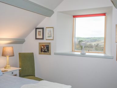 A bedroom corner with a green chair next to a white nightstand with a lamp and framed pictures on the wall at Hen Beudy in Porthmadog