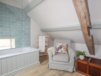 A bathroom with a bathtub wooden towel rack armchair with patterned cushion wooden chest with a plant and exposed wooden beam at Hen Beudy in Porthmadog