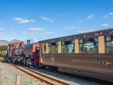 A vintage steam train pulling a passenger car labeled Bodysgallen on tracks with mountains in the background at Hen Beudy in Porthmadog