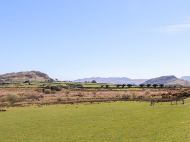 A grassy field with trees and hills in the background under a clear blue sky at Hen Beudy in Porthmadog