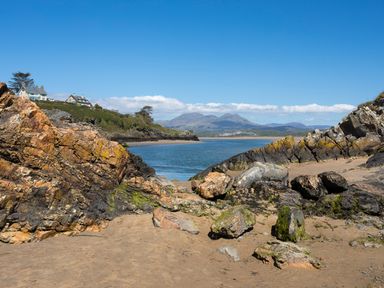 A rocky beach with sand and water with houses and mountains in the background at Hen Beudy in Porthmadog