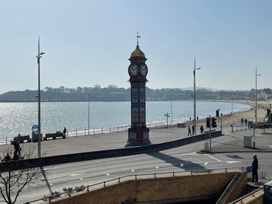 A clock tower by a waterfront with a beach and people walking near Sunrise Apartment in Weymouth