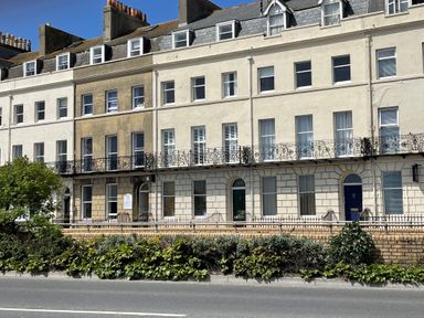 A row of multi-story buildings with balconies and windows along a street with greenery in front at Sunrise Apartment in Weymouth