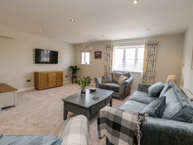 A living room with grey sofas a coffee table a wooden cabinet a wall-mounted TV and windows with patterned curtains at Rose Cottage in Faceby near Swainby