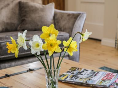 A vase with yellow and white daffodils on a wooden table with magazines and a grey sofa in the background at Rose Cottage Faceby near Swainby