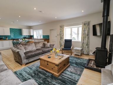 An open living room and kitchen with sofas a wooden coffee table a blue armchair a wood stove and turquoise tiled kitchen at Rose Cottage Faceby near Swainby