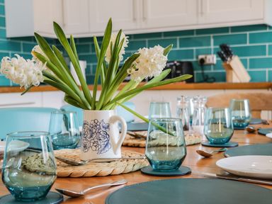 A dining table set with blue glasses white plates and a white jug with flowers in a kitchen with teal tiled backsplash at Rose Cottage in Faceby near Swainby