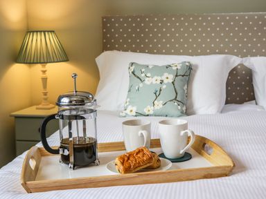A breakfast tray with coffee pot two cups and a pastry on a bed with a polka dot headboard and floral cushion at Rose Cottage in Faceby near Swainby