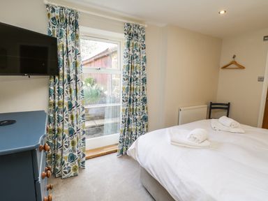A bedroom with a bed with white linens and towels a blue dresser with wooden knobs a wall-mounted TV floral curtains and a window at Rose Cottage in Faceby near Swainby