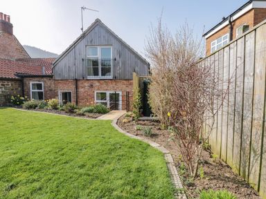 A backyard garden with grass lawn and flower beds next to a brick and wood house at Rose Cottage in Faceby near Swainby