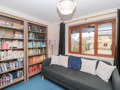 A room with bookshelves filled with books and DVDs a gray sofa with white and blue cushions a window with a view of houses and a lamp next to the sofa at Morgan House in Middlezoy