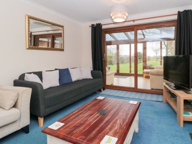 A living room with a dark sofa and cushions a wooden coffee table a TV on a wooden stand and glass sliding doors leading to a conservatory at Morgan House in Middlezoy