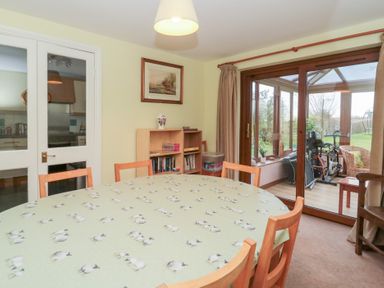 A dining room with a round table covered with a sheep-patterned tablecloth surrounded by wooden chairs a shelving unit with books and a vase a framed picture on the wall glass doors leading to a conservatory with exercise equipment and garden view at Morgan House in Middlezoy