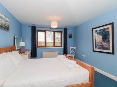 A bedroom with a wooden bed frame white bedding two rolled towels a window with dark curtains a white bedside table with a lamp a white chair and framed artwork on blue walls at Morgan House in Middlezoy