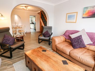 A living room with a brown leather sofa and two gray chairs around a wooden coffee table with a dining area and kitchen visible through an arch at 57 Southend Road Hunstanton