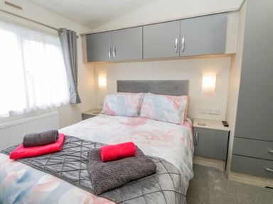 A bedroom with a double bed floral bedding overhead cabinets and folded towels at Happy Daze Carnaby near Bridlington