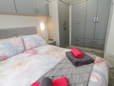 A bedroom with a bed covered in floral bedding and folded towels on top with wardrobes and cabinets along the walls at Happy Daze in Carnaby near Bridlington