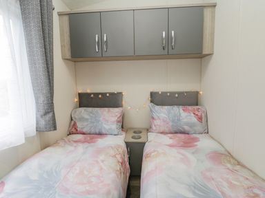 A bedroom with two single beds with floral bedding a small nightstand with built in power outlets overhead cupboards and a window with patterned curtains at Happy Daze in Carnaby near Bridlington