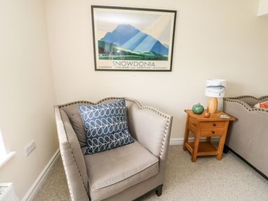 A corner chair with cushions next to a wooden side table with a lamp and decoration under a snowdonia framed picture at 8 Llwyn Onn in Llanfairpwllgwyngyll