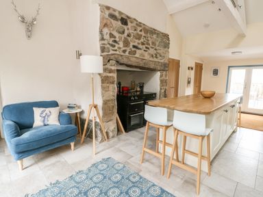 A kitchen with a stone fireplace a blue armchair a wooden island with two stools and a patterned rug at 8 Llwyn Onn in Llanfairpwllgwyngyll