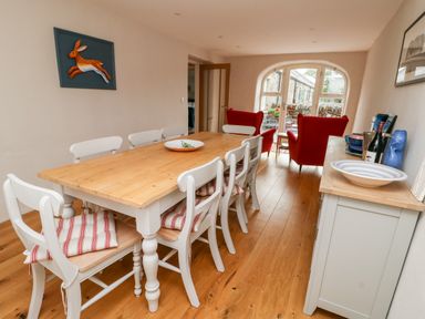 A dining room with a wooden table and white chairs with cushions and red armchairs at 8 Llwyn Onn in Llanfairpwllgwyngyll