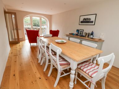 A dining room with a wooden table and white chairs with cushions and red armchairs near a window at 8 Llwyn Onn in Llanfairpwllgwyngyll