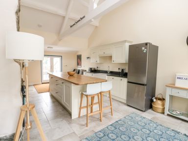A kitchen with a wooden island and two white stools a stainless steel refrigerator a floor lamp and a blue patterned rug at 8 Llwyn Onn in Llanfairpwllgwyngyll