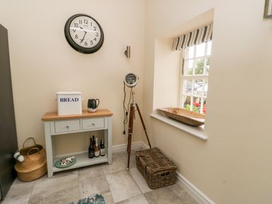 A corner with a small table holding a bread box and jug a large clock on the wall a tripod lamp a wicker basket and a window with a wooden bowl on the sill at 8 Llwyn Onn in Llanfairpwllgwyngyll
