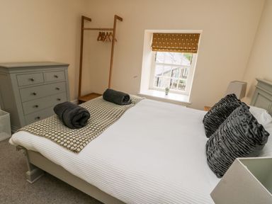 A bedroom with a bed made with white bedding and dark pillows beside a window with a patterned blind and a gray dresser at 8 Llwyn Onn in Llanfairpwllgwyngyll