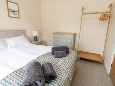 A bedroom with a bed with rolled towels a bedside table with lamp a chest of drawers and a wooden clothes rack at 8 Llwyn Onn in Llanfairpwllgwyngyll