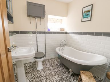 A bathroom with a pedestal sink, toilet, clawfoot bathtub, patterned floor tiles, and a small wooden stool at 8 Llwyn Onn in Llanfairpwllgwyngyll