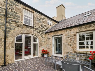 A stone patio with metal chairs and a table outside a stone house with arched windows and red flowers at 8 Llwyn Onn in Llanfairpwllgwyngyll