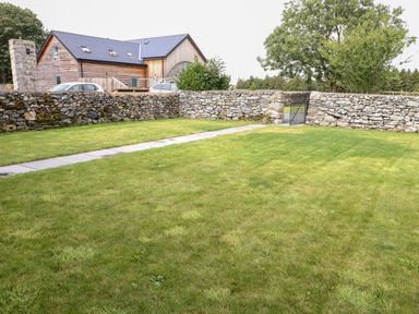 A grassy yard with a stone wall and a gate near a wooden house with cars parked at 8 Llwyn Onn in Llanfairpwllgwyngyll