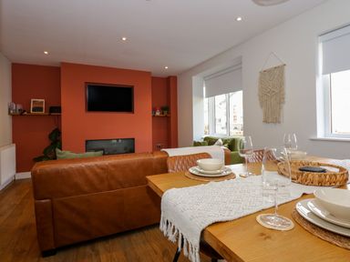 A dining table set with plates, bowls, and wine glasses next to brown leather sofas facing a wall-mounted TV above a fireplace at Aran in Beddgelert