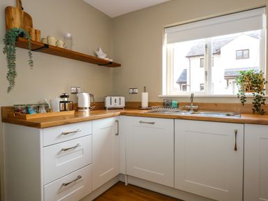 A kitchen corner with white cabinets wooden countertop a sink window with a blind a floating wooden shelf with mugs plates and decor at Aran in Beddgelert