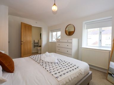 A bedroom with a bed with folded towels a white chest of drawers a round mirror two windows and an open door leading to a bathroom at Aran in Beddgelert
