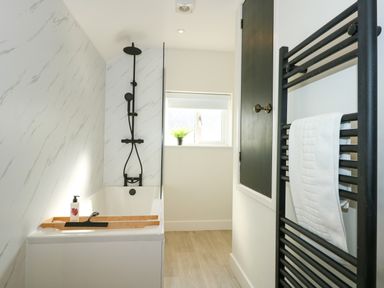 A bathroom with a bathtub featuring a black showerhead and faucet a wooden tray with soap on the tub a towel hanging on a black heated towel rail a small window with a plant on the windowsill at Aran in Beddgelert