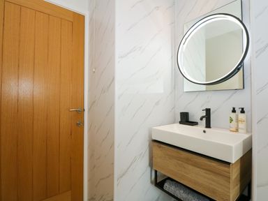 A bathroom with a wooden door a wall-mounted sink with toiletries and a round illuminated mirror at Aran in Beddgelert