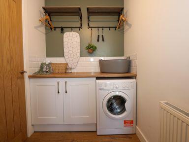 A laundry room with a washing machine white cabinets wooden countertop ironing board and shelves at Craig y Llan Beddgelert