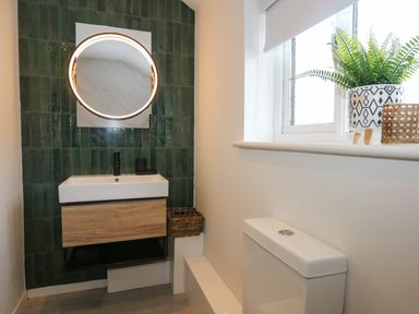 A bathroom with a sink cabinet and round mirror on a green tiled wall and a toilet under a window with a plant at Craig y Llan in Beddgelert