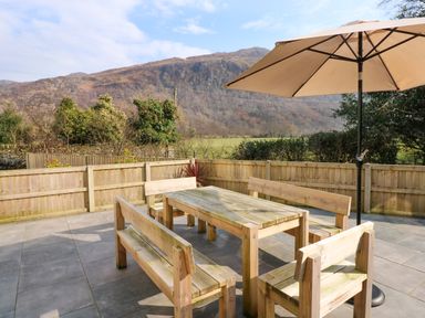 An outdoor patio with wooden table and benches under a large umbrella with mountains and trees in the background at Craig y Llan in Beddgelert