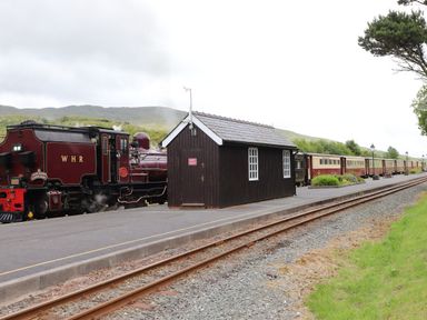 A steam train stopped at a rural train station platform with a small wooden building and railway tracks at Craig y Llan in Beddgelert